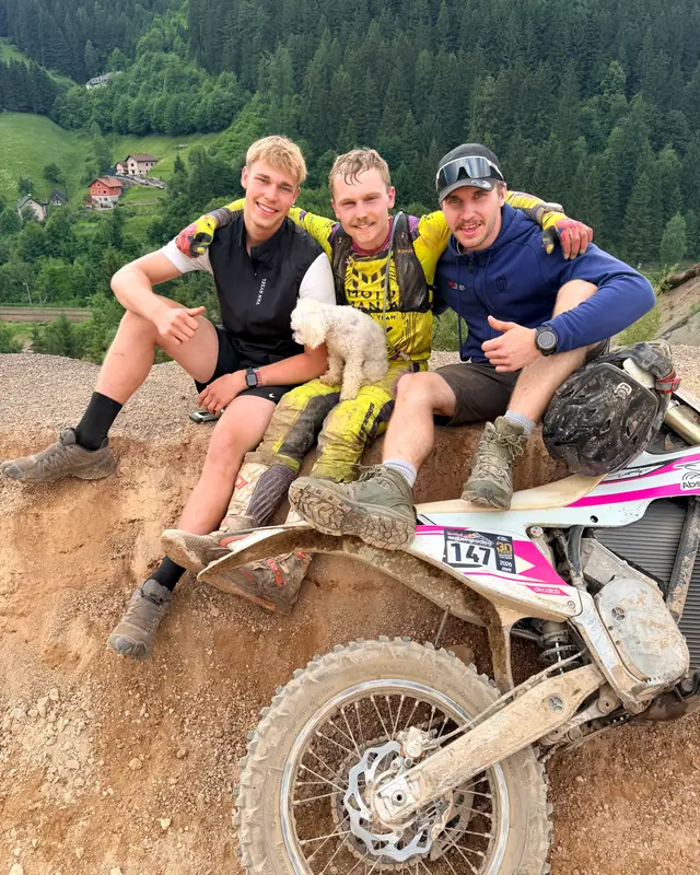 Three men with a small dog sit on a mountain slope next to a motorcycle, forested mountains behind