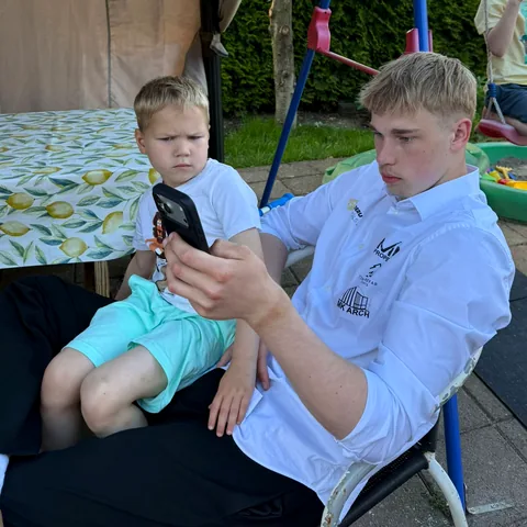 Gustavs baltā kreklā ar telefonu rokā, pagalmā